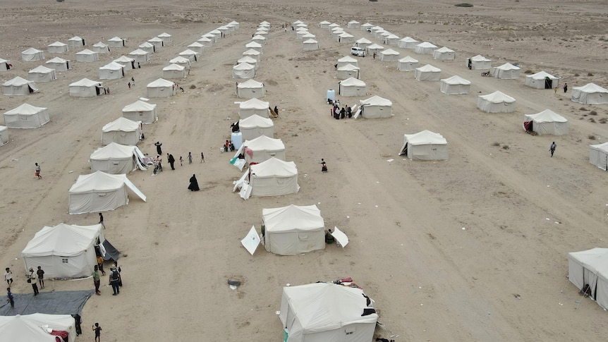 A drone image of a camp consisting of dozens of white tents standing in the desert, people can be seen on the ground