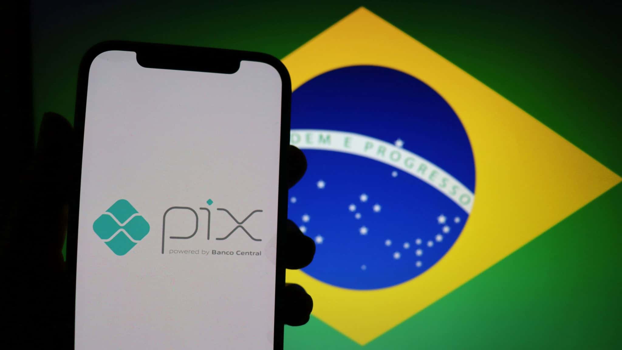 Pessoa segura celular exibindo logomarca do Pix na tela; ao fundo, monitor exibe bandeira do Brasil